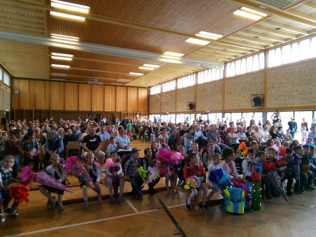 Eine große Gruppe kleiner Kinder sitzt mit bunten Blumensträußen in der Hand vor einer überfüllten Schulturnhalle, dahinter sitzen viele Erwachsene und Eltern und schauen sich eine Veranstaltung an.