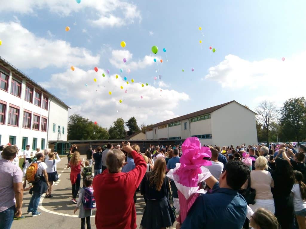 Eine große Gruppe von Menschen hat sich auf einem Schulhof versammelt und beobachtet und fotografiert, wie an einem sonnigen Tag bunte Ballons in den Himmel steigen. Einige Kinder und Erwachsene halten Blumen und Rucksäcke in den Händen.