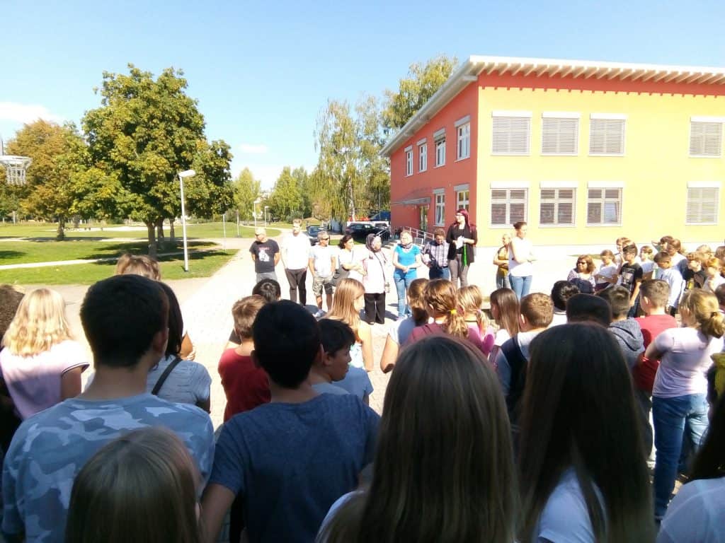 Eine große Gruppe von Schülern steht an einem sonnigen Tag vor einem roten Gebäude mit großen Fenstern, während einige Erwachsene mit ihnen sprechen. Im Hintergrund sind Bäume und Gras zu sehen.