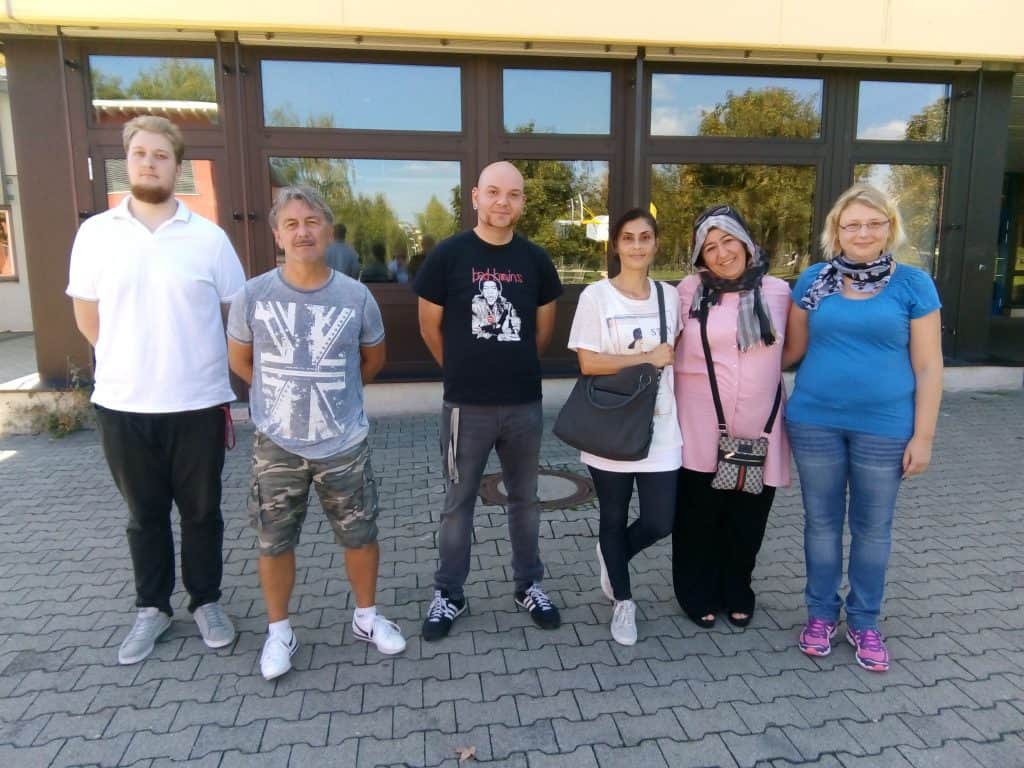 Sechs Erwachsene stehen nebeneinander auf einer gepflasterten Fläche vor einem Gebäude mit großen Fenstern und lächeln in die Kamera. Die Gruppe besteht aus drei Männern und drei Frauen in Freizeitkleidung.