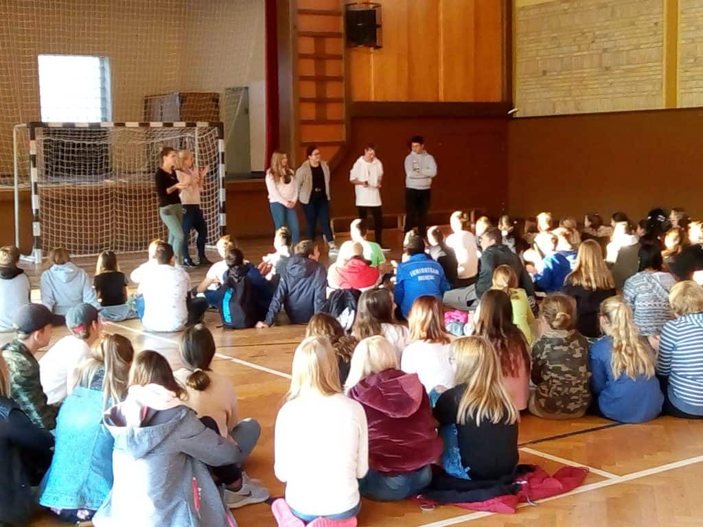 Eine Gruppe von Schülern sitzt in einer Turnhalle auf dem Boden, gegenüber einigen Personen, die in der Nähe eines Tornetzes stehen und sich unterhalten. Es erinnert an eine Schulversammlung oder Präsentation. Sonnenlicht fällt durch die Fenster auf der linken Seite.