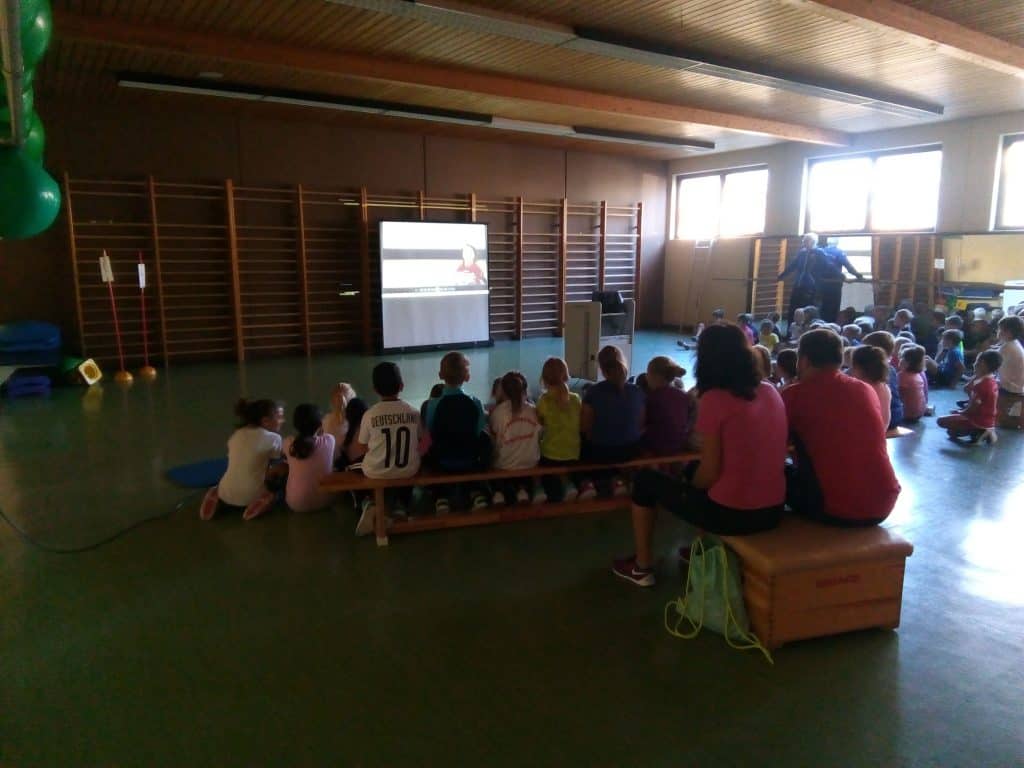 Kinder sitzen auf Bänken in einer Turnhalle und schauen sich ein Video auf einer Leinwand an. Der Raum ist schwach beleuchtet, und einige Erwachsene sind in der Gruppe. An den Wänden sind Turngeräte zu sehen.