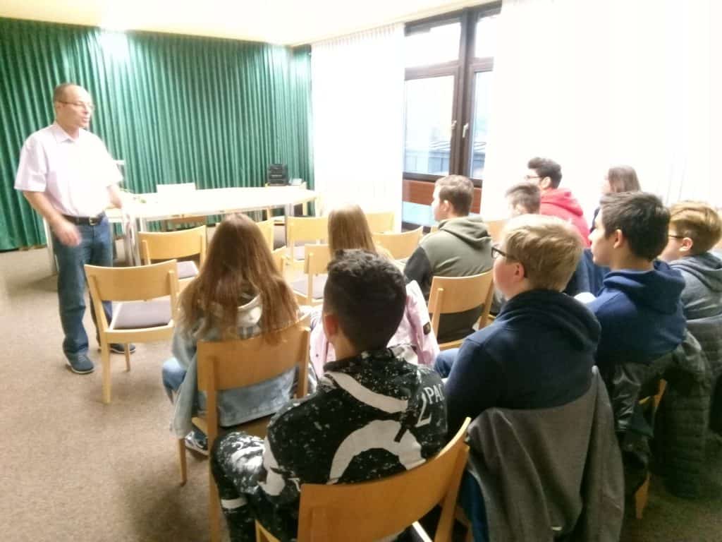 Ein Mann steht vor einem Raum und spricht mit einer Gruppe sitzender Teenager, die aufmerksam zuhören. Der Raum hat grüne Vorhänge, große Fenster und mehrere in Reihen angeordnete Holzstühle.