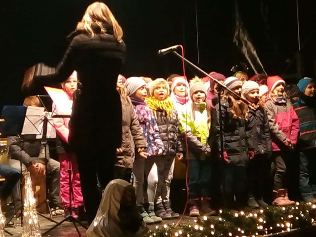 Eine Gruppe von Kindern in Winterkleidung singt nachts unter der Leitung eines Dirigenten auf der Bühne. Sie stehen vor einem Mikrofon, umgeben von festlichen Lichtern und Dekorationen.