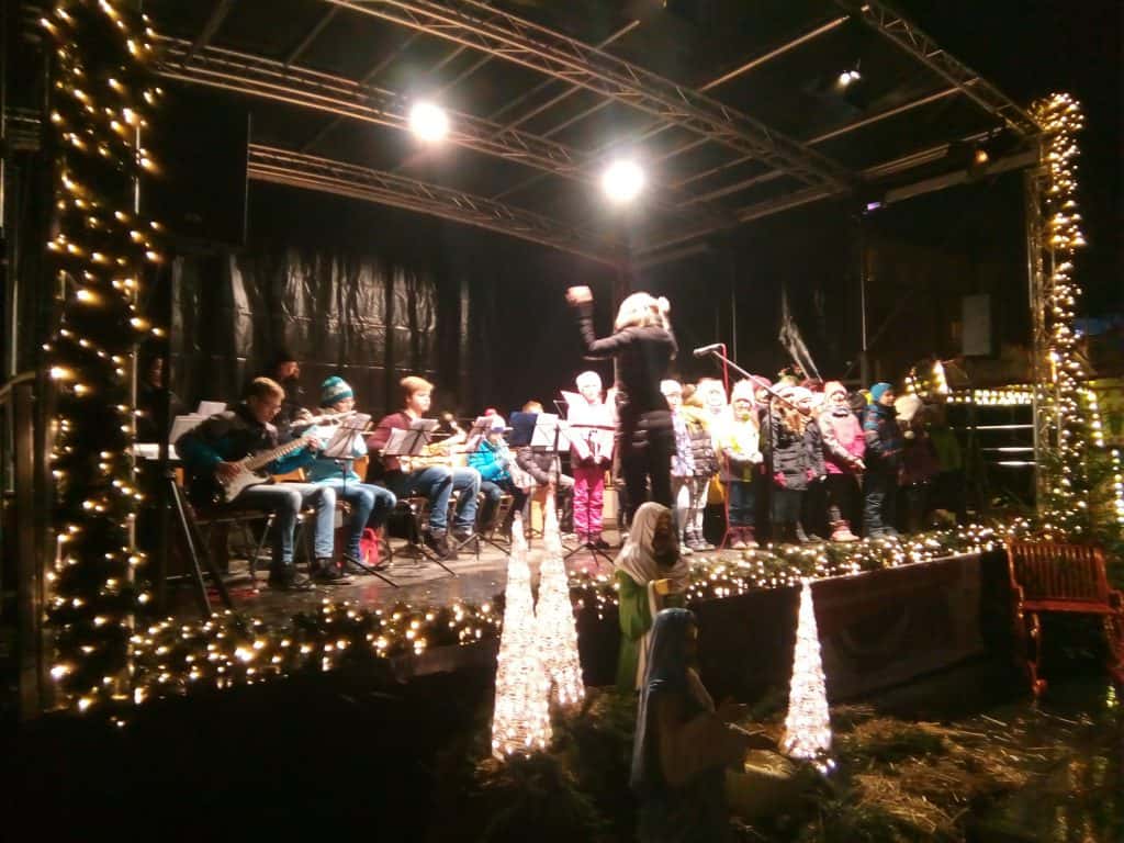 Ein Dirigent leitet eine Gruppe singender Kinder auf einer mit Weihnachtslichtern und Weihnachtsbäumen geschmückten Bühne. Links spielen Musiker Instrumente, und im Vordergrund sind Krippenfiguren zu sehen.