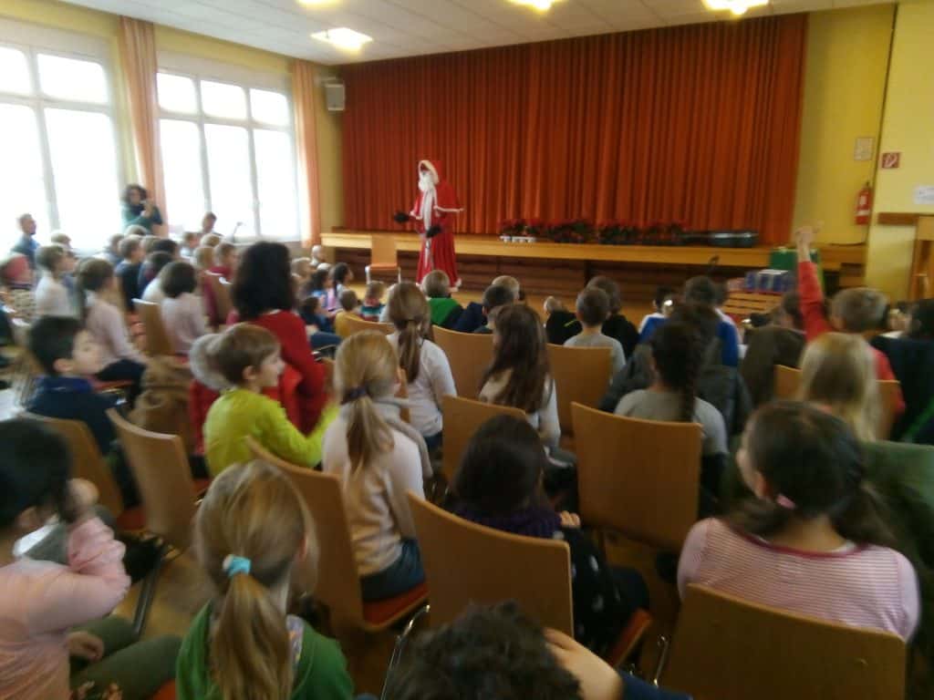 Eine Frau in einem rot-weißen Kostüm steht auf einer Bühne vor einem sitzenden Kinderpublikum in einem hell erleuchteten Raum mit großen Fenstern und orangefarbenen Vorhängen.