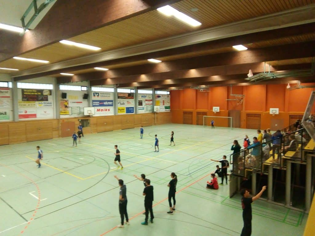 In einer großen Sporthalle läuft ein Basketballspiel. Die Spieler tragen blau-schwarze Trikots. Zuschauer sitzen und stehen auf den Tribünen rechts. An den Wänden hängen Werbebanner.