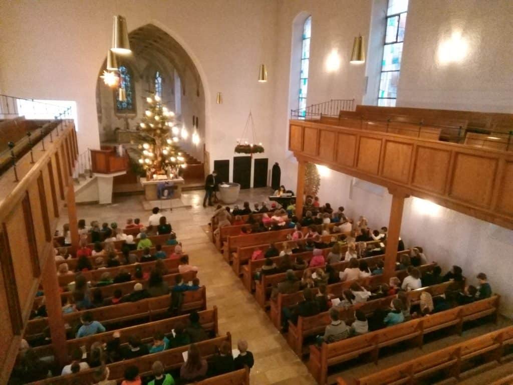 Ein Blick von oben zeigt Menschen in den Kirchenbänken einer Kirche mit hohen Decken, großen Fenstern und einem geschmückten Weihnachtsbaum in der Nähe des Altars. Die Kirche ist mit Hängelampen gut beleuchtet.