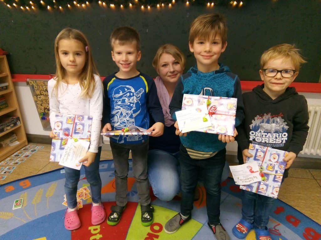 Eine Frau kniet hinter vier lächelnden Kindern, die bunte Umschläge und Karten halten. Sie stehen auf einem Klassenzimmerteppich, im Hintergrund sind Weihnachtsdekorationen und Lichter zu sehen.