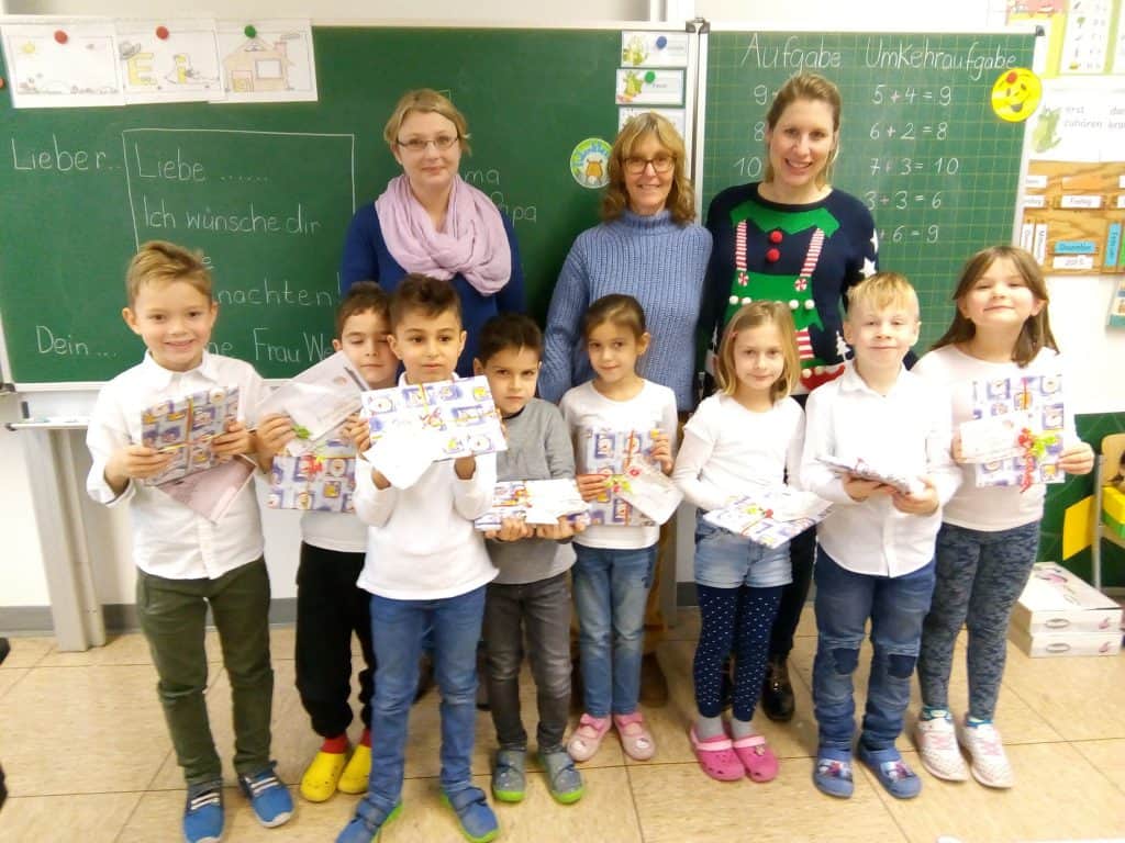 Eine Gruppe von acht Kindern mit Büchern und Papieren steht mit drei erwachsenen Frauen in einem Klassenzimmer vor einer Tafel. Die Kinder lächeln, und die Erwachsenen stehen hinter ihnen und lächeln ebenfalls.