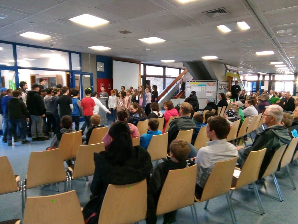 Eine Gruppe von Kindern steht vor einem Publikum, das auf Holzstühlen in einem hell erleuchteten Innenraum sitzt, möglicherweise einer Schule oder einem Gemeindezentrum. Im Hintergrund sind Plakate und Treppen zu sehen.