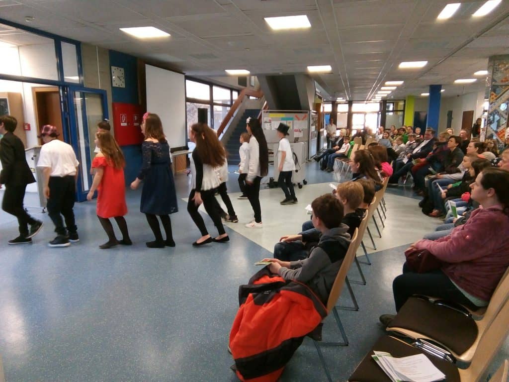 Eine Gruppe von Kindern führt in einem Schulflur einen Tanz oder ein Theaterstück auf, während ein Publikum aus Erwachsenen und Kindern aufmerksam zusieht. Im Hintergrund sind Klassenzimmertüren und Anzeigetafeln zu sehen.