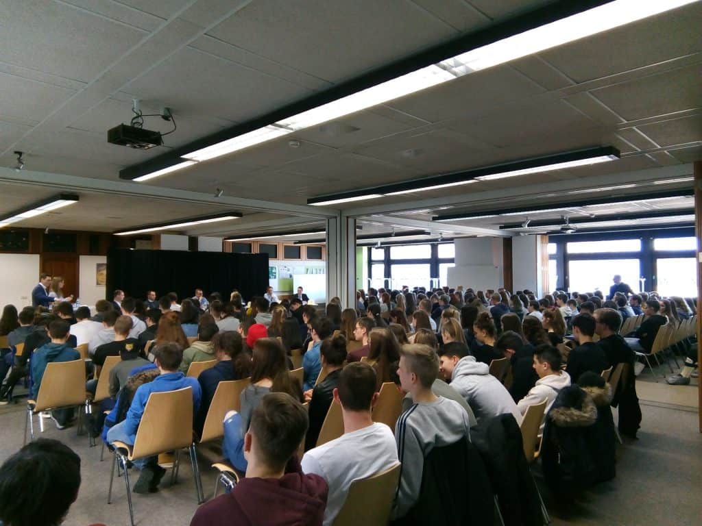 Eine große Gruppe von Menschen, hauptsächlich junge Erwachsene, sitzt auf Stühlen vor einer Bühne in einem hellen, geräumigen Raum mit großen Fenstern und Neonlicht und hört einer Präsentation oder Vorlesung zu.