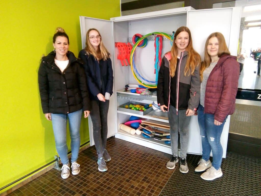 Vier Mädchen stehen lächelnd vor einem offenen weißen Schrank voller bunter Sport- und Spielgeräte wie Reifen und Bälle vor einer hellgrünen Wand im Innenbereich.