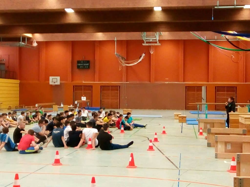 Eine Gruppe von Schülern sitzt in einer Turnhalle auf dem Boden und hört einem Trainer zu. Die Halle hat orangefarbene Wände, Sportgeräte und orangefarbene Kegel, die die Bereiche markieren. Im Hintergrund sind hölzerne Sprungkästen und Matten zu sehen.