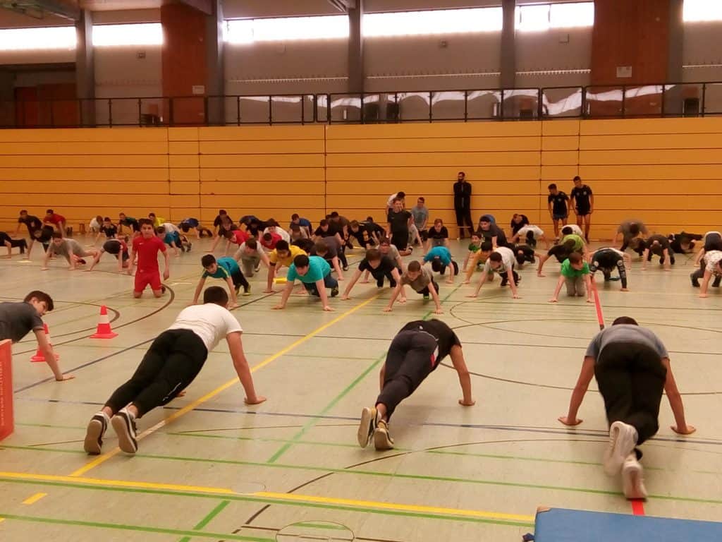 Eine Gruppe von Menschen in Sportkleidung macht Liegestütze auf dem Boden einer großen Turnhalle mit gelben Wänden und mehreren Zuschauern im Hintergrund.