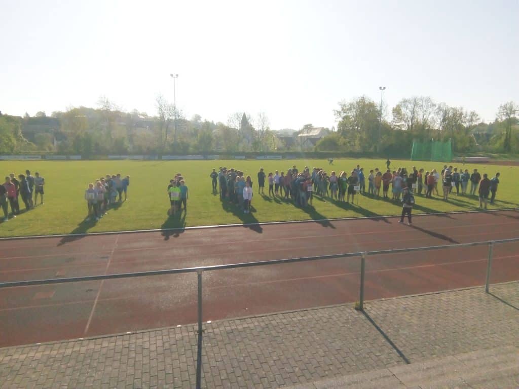 Gruppen von Menschen stehen mit Abstand auf einer Wiese in der Nähe einer Laufbahn im hellen Sonnenlicht. Einige haben sich in Gruppen versammelt, andere sind verstreut, mit Bäumen und Gebäuden im Hintergrund.