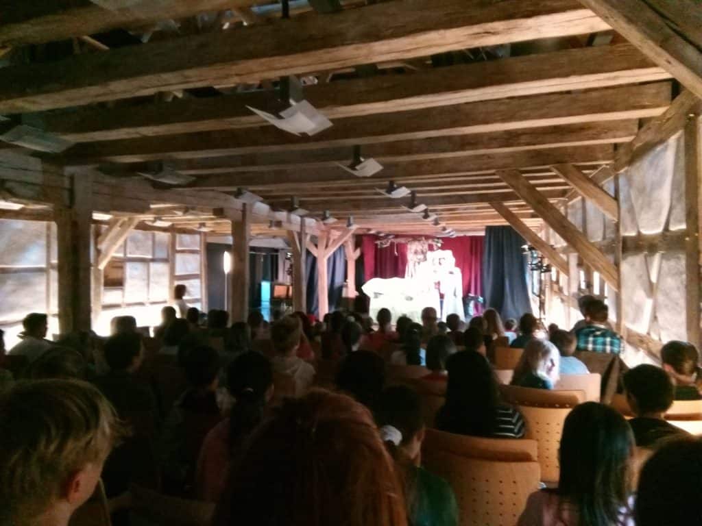 Das Publikum sitzt in einem rustikalen Holzsaal und blickt auf eine hell erleuchtete Bühne mit Künstlern im Vordergrund. Der Raum ist voller Menschen, und darüber sind die Balken der Holzdecke sichtbar.