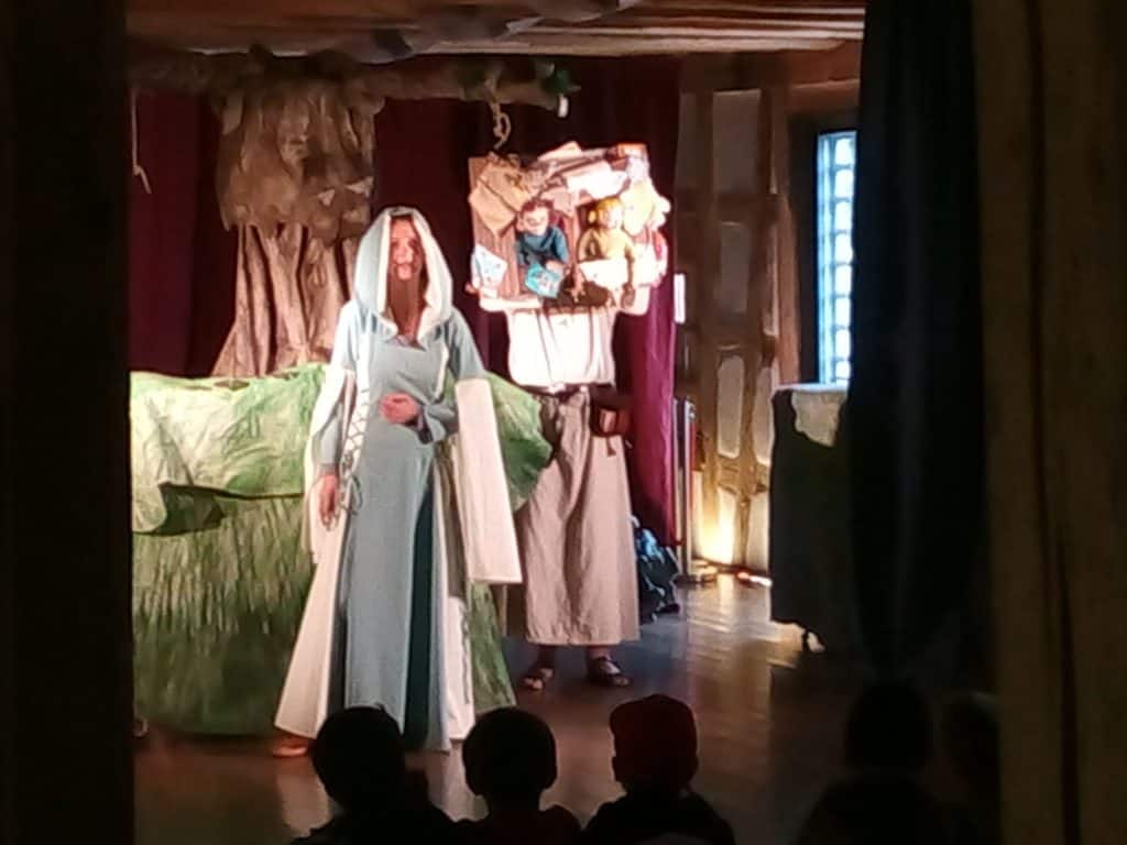 Eine Frau in einem hellblauen Gewand und Schleier steht lächelnd auf der Bühne. Hinter ihr hält eine kostümierte Person eine bunte Collage. Das Bühnenbild umfasst einen großen Baum und eine grüne Landschaft. Kinder im Publikum schauen zu.