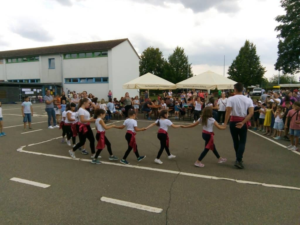 Eine Gruppe von Kindern und ein Erwachsener, alle in weißen Hemden und roten Schärpen, halten sich an den Händen und tanzen im Freien im Kreis, während eine Menschenmenge zuschaut. Die Veranstaltung findet auf einem Spielplatz in der Nähe eines Gebäudes statt.