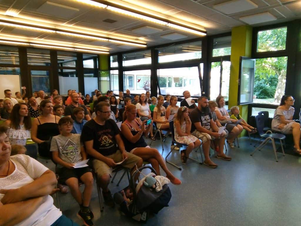 Eine Gruppe von Erwachsenen und Kindern sitzt in einem hellen Klassenzimmer auf Stühlen, blickt nach vorne und hört aufmerksam zu; große Fenster lassen natürliches Licht herein und einige Leute halten Papiere oder Telefone in den Händen.