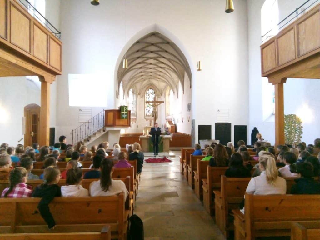 Eine Gruppe von Menschen sitzt auf Holzbänken in einer hellen Kirche und blickt auf einen Redner am Altar. Die Kirche hat hohe Decken, hohe Bogenfenster und ein großes Kreuz an der Vorderseite.