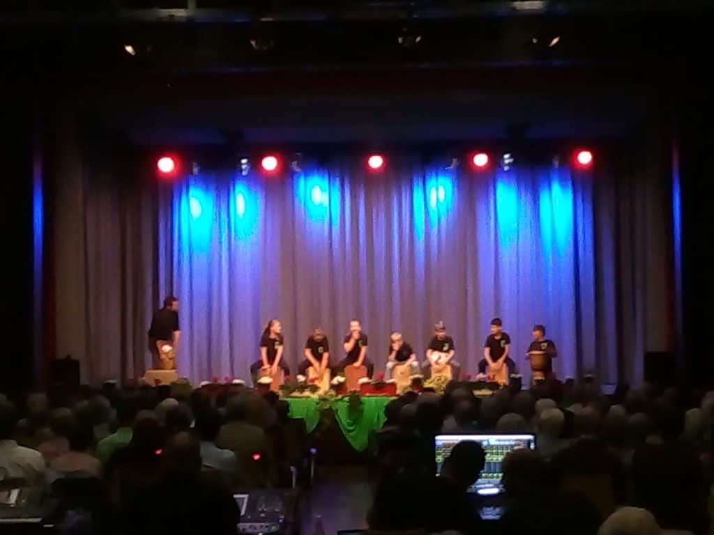 Eine Gruppe von Menschen tritt mit Trommeln und Schlaginstrumenten unter blauem und rotem Licht auf der Bühne auf und steht einem Publikum in einem dunklen Zuschauerraum gegenüber.