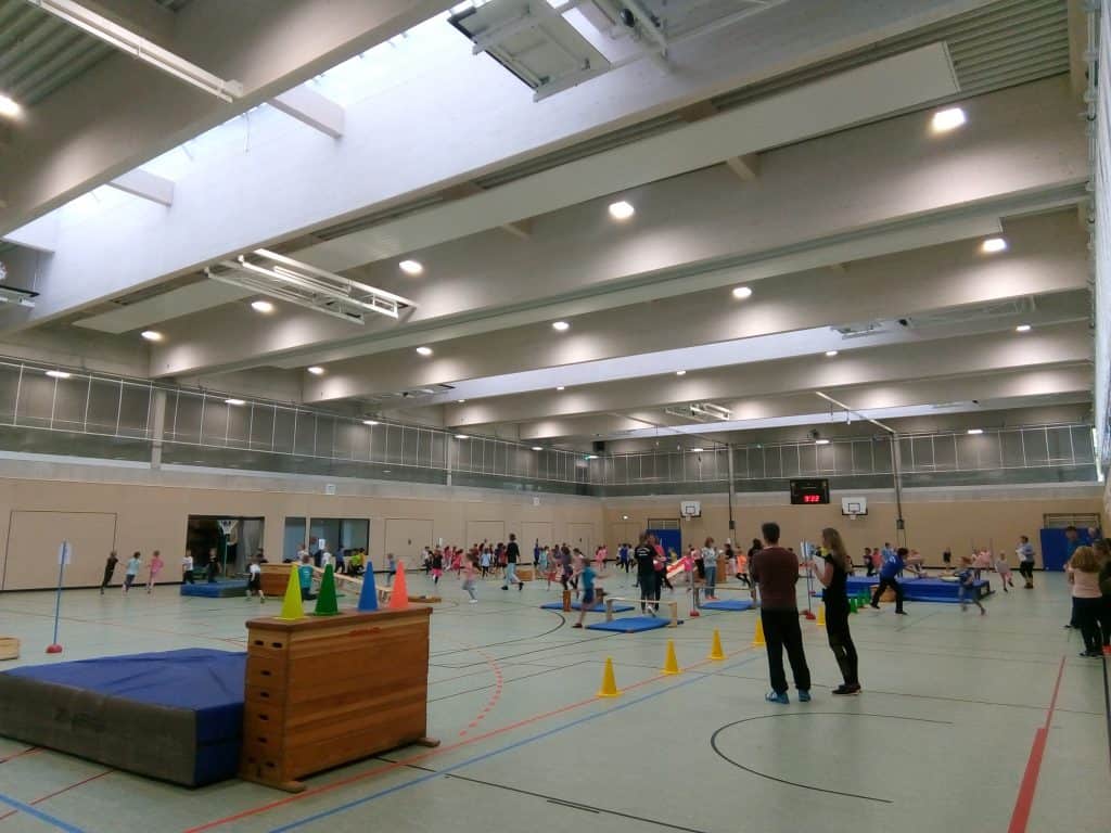 Kinder und Erwachsene nehmen an Aktivitäten in einer geräumigen Turnhalle mit hohen Decken, Sportgeräten und bunten Kegeln teil. Einige Menschen haben sich in der Nähe von Matten und Turngeräten versammelt.