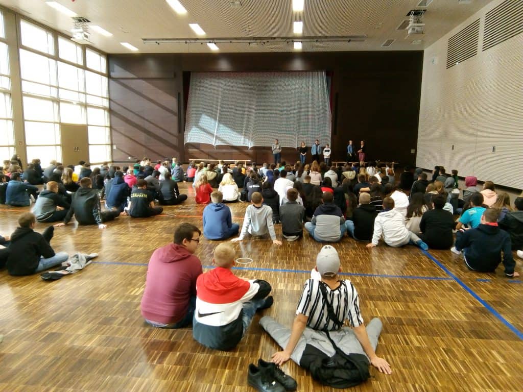 Eine große Gruppe von Studierenden sitzt auf dem Boden einer Turnhalle vor einer Bühne, auf der mehrere Personen vor einem hochgezogenen Vorhang stehen und sprechen. Der Raum ist hell erleuchtet durch Tageslicht, das durch hohe Fenster hereinfällt.