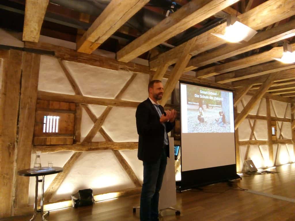 Ein Mann steht und spricht vor einer Projektionsleinwand, auf der „Smart School: Die Schule der Zukunft“ läuft. Die Leinwand befindet sich in einem rustikalen Raum mit freiliegenden Holzbalken und einem Laptop auf einem Tisch in der Nähe.