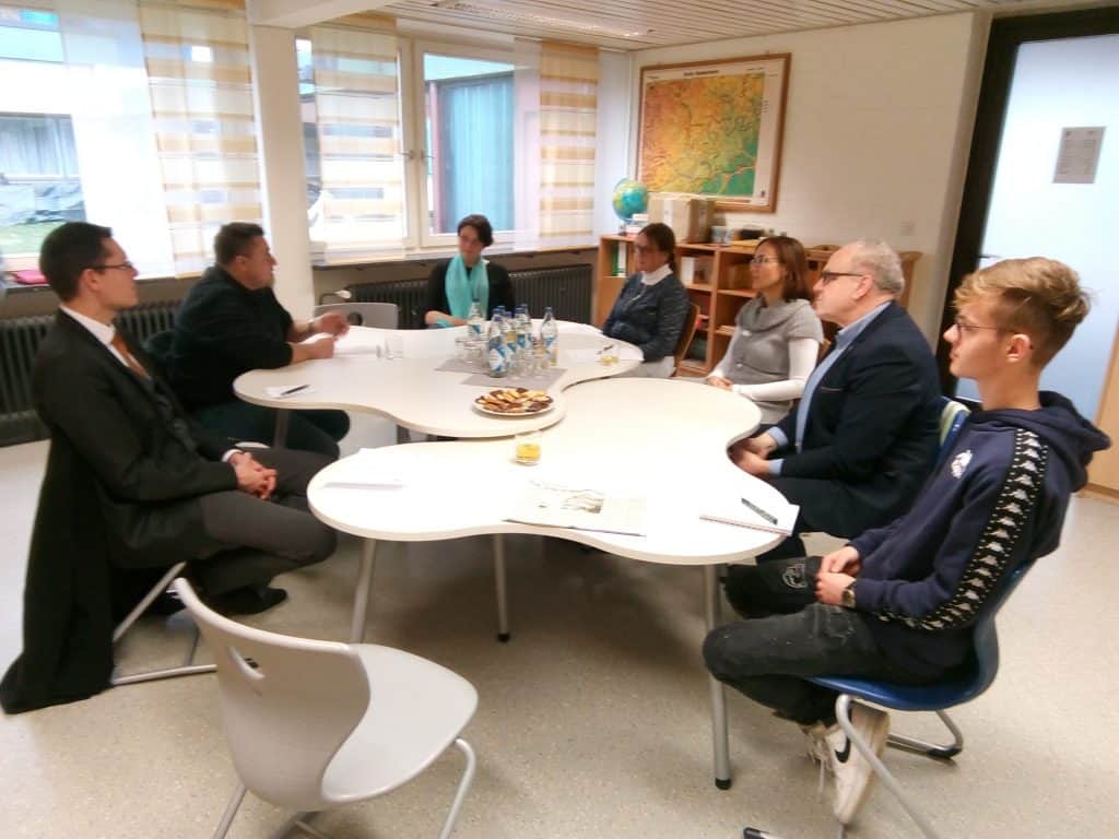 Sieben Personen sitzen in einem Klassenzimmer um einen weißen Tisch und diskutieren. Auf dem Tisch stehen Papiere, Snacks und Getränke. Im Hintergrund sind neben den Fenstern eine Karte und ein Globus zu sehen.