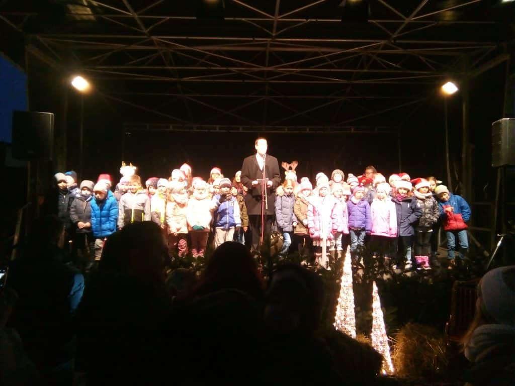 Eine Gruppe Kinder in Winterkleidung und mit Mützen steht auf einer Freilichtbühne. Ein Mann hält ein Mikrofon. Die Bühne ist mit Weihnachtslichtern und Weihnachtsbäumen geschmückt. Im Vordergrund ist ein Publikum zu sehen.