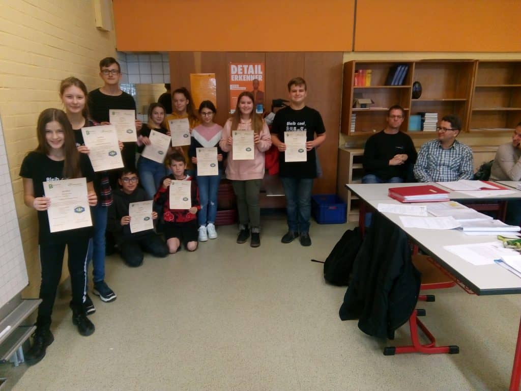 Eine Gruppe von Schülern steht mit Zertifikaten in einem Klassenzimmer und lächelt in die Kamera. Rechts sitzen mehrere Erwachsene an den Tischen und beobachten das Geschehen. Im Hintergrund sind Regale und Unterrichtsmaterialien zu sehen.