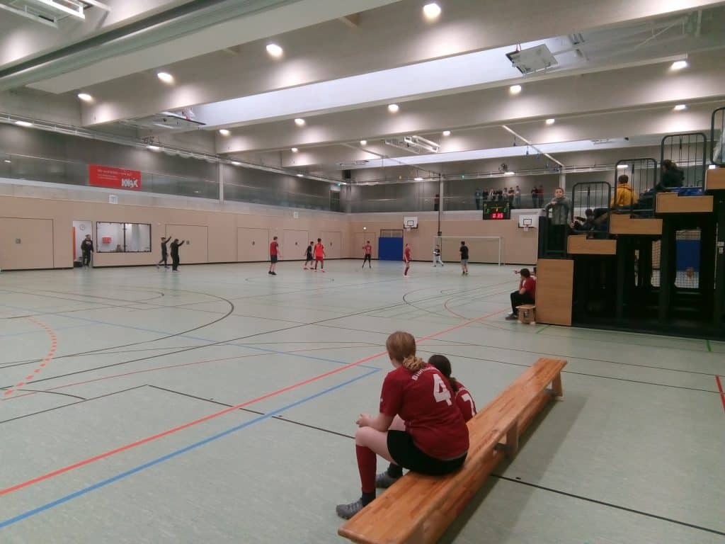 Sporthalle mit einer Gruppe von Menschen, die ein Spiel spielen, möglicherweise Futsal oder Handball. Zwei Spieler in roten Trikots sitzen im Vordergrund auf einer Bank. Zuschauer sitzen auf erhöhten Tribünen rechts.