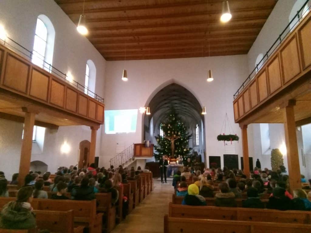 Eine große Gruppe von Menschen sitzt in Holzbänken in einer Kirche mit hohen Decken, vorn steht ein geschmückter Weihnachtsbaum und in der Nähe des Altars hält ein Redner eine Ansprache an das Publikum.