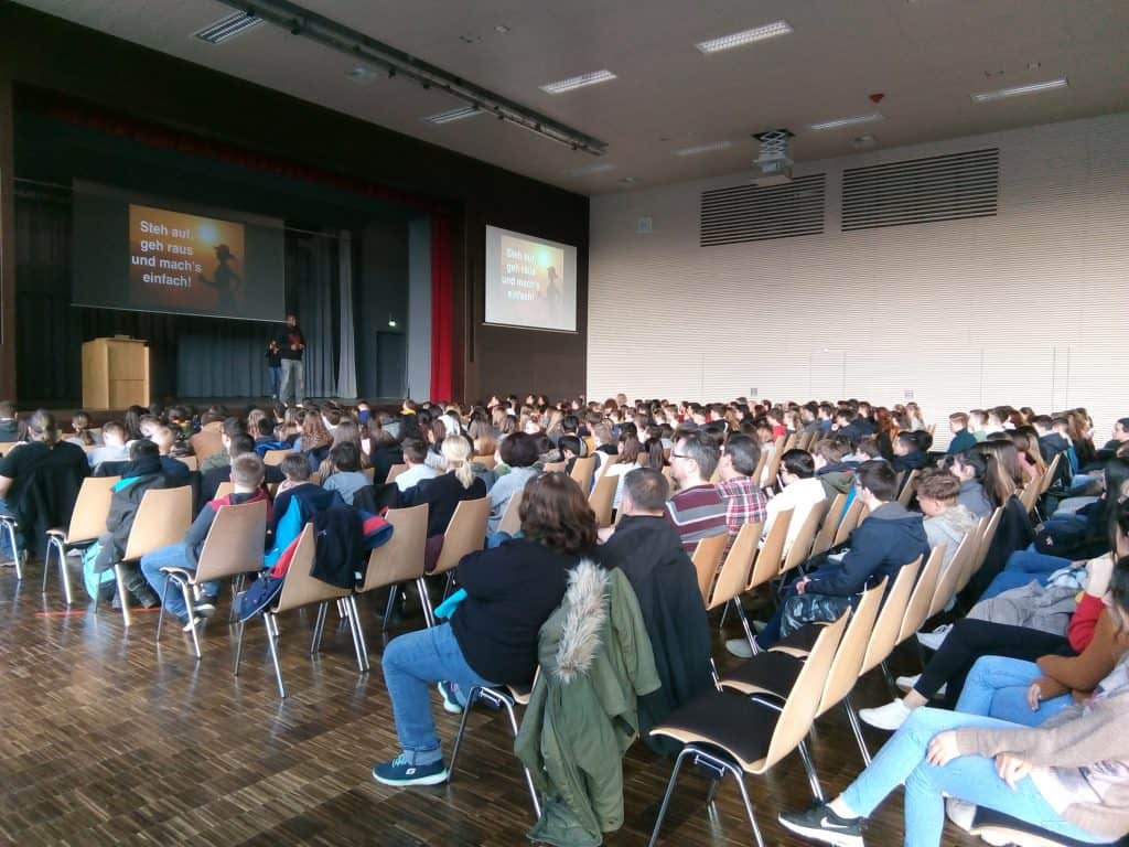 Eine große Gruppe von Menschen sitzt auf Stühlen in einem Auditorium vor einer Bühne, auf der ein Redner einen Vortrag hält. Auf einem Bildschirm wird deutscher Text angezeigt. Der Raum hat Holzböden, hohe Decken und Tageslicht.