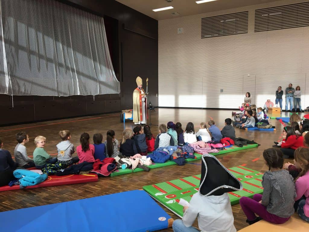 Eine als Nikolaus verkleidete Person steht auf einer Bühne und spricht zu einer Gruppe von Kindern, die in einem großen, hellen Raum auf Matten sitzen. Einige Kinder tragen Kostüme, andere schauen aufmerksam zu.