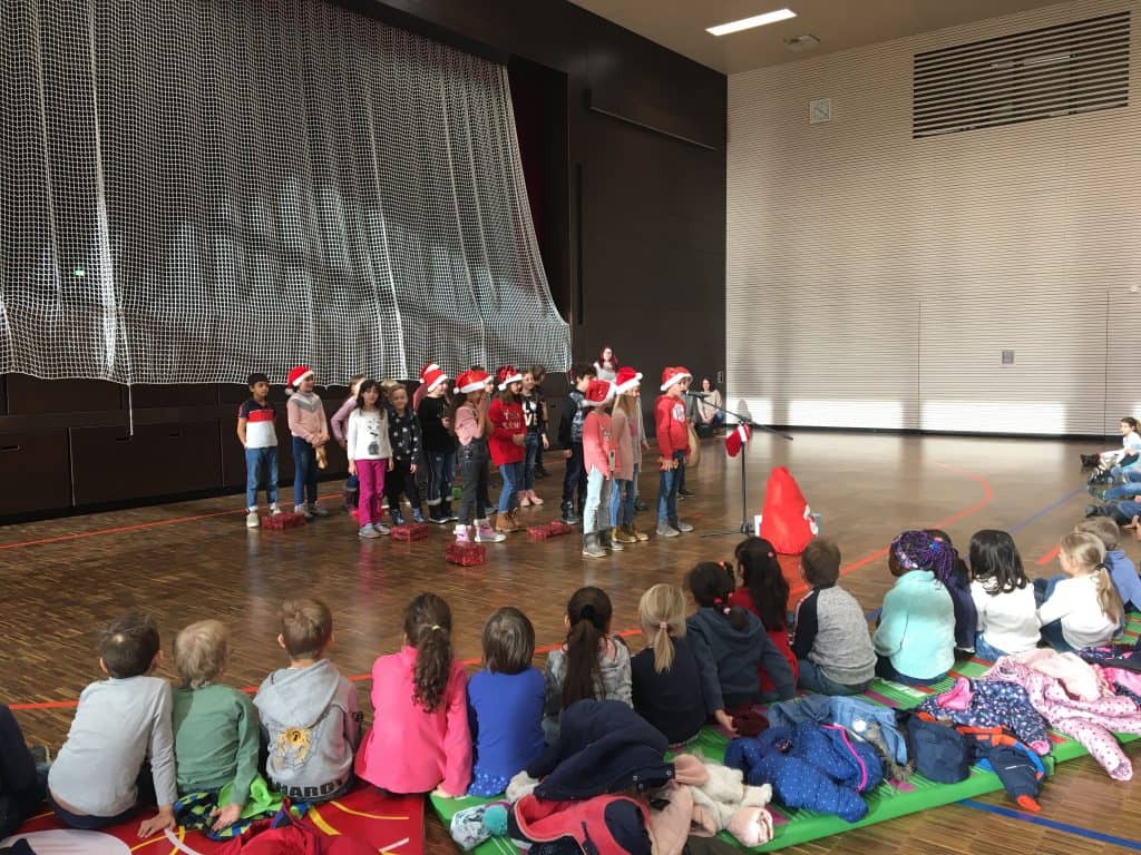 Eine Gruppe von Kindern mit Weihnachtsmannmützen tritt auf einer Bühne in einer Turnhalle auf, während ein Publikum aus sitzenden Kindern zusieht. Sonnenlicht fällt herein, und an der Rückwand hängt ein großes Netz.