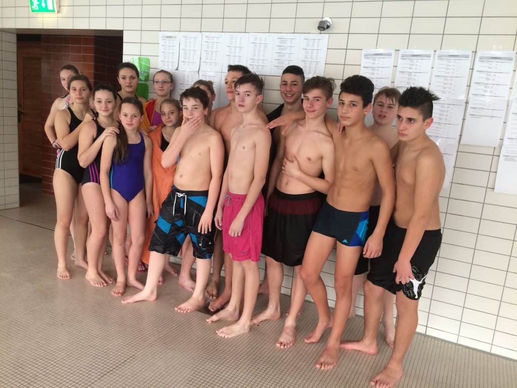 Eine Gruppe Teenager in Badeanzügen steht zusammen in einem Gebäude an einer gefliesten Wand, an der Zettel hängen, und posiert für ein Gruppenfoto in einer Anlage, die wie ein Schwimmbad aussieht.