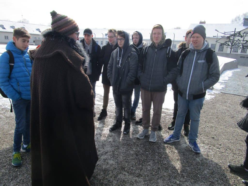 Eine Gruppe junger Leute in Winterkleidung steht an einem verschneiten Tag im Freien und hört einer Person in braunem Mantel und Hut zu. Im Hintergrund sind Gebäude zu sehen.