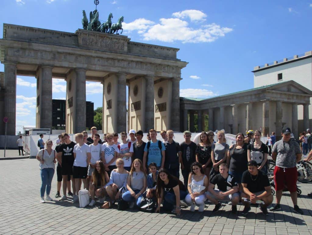 Eine große Gruppe junger Leute posiert an einem sonnigen Tag mit blauem Himmel und vereinzelten Wolken für ein Foto vor dem Brandenburger Tor in Berlin.