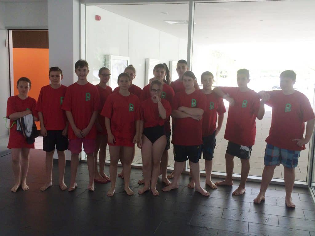 Eine Gruppe von dreizehn jungen Leuten in passenden roten Hemden und Badeanzügen steht in einem Innenbereich auf einem dunklen Boden und posiert gemeinsam vor großen Glasfenstern.