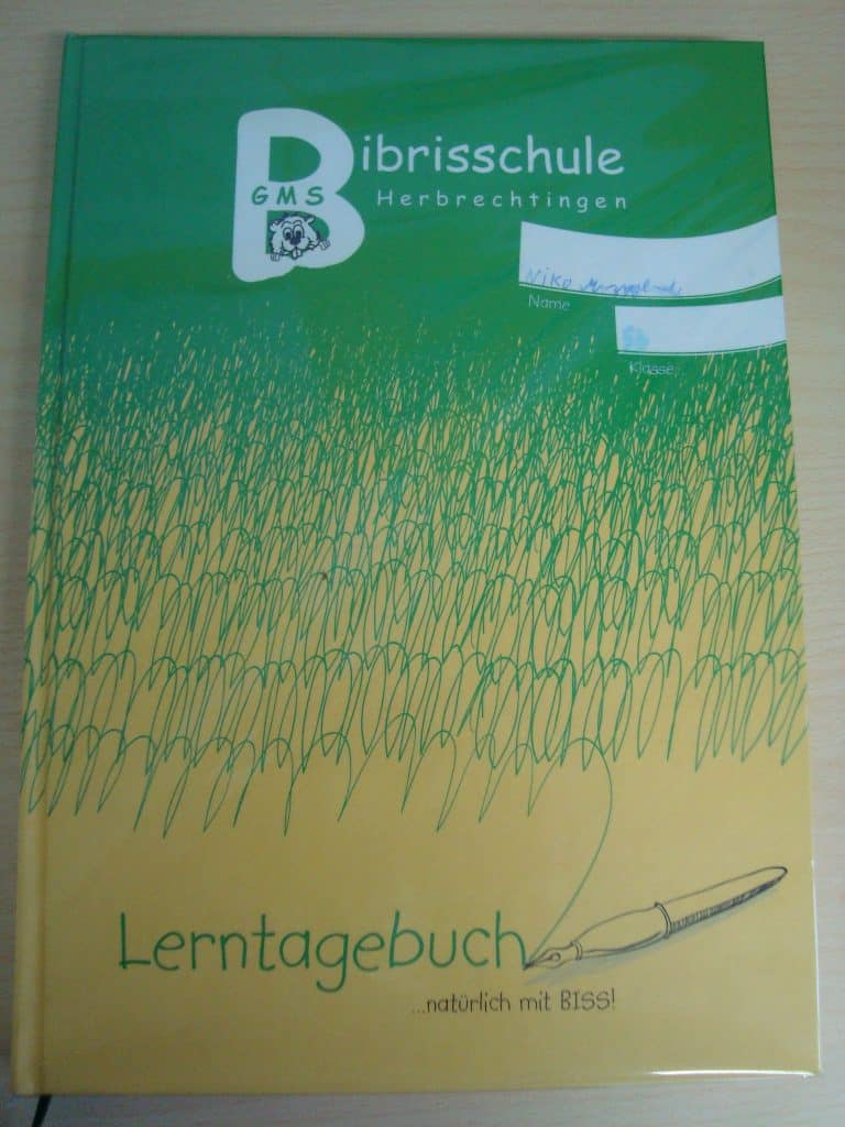 Ein grün-gelbes Schulheft mit dem Titel „Lerntagebuch“ der Bibrisschule Herbrechtingen, mit einer Grasillustration und einem Stift. Oben rechts befindet sich ein Etikett mit einem handgeschriebenen Namen.