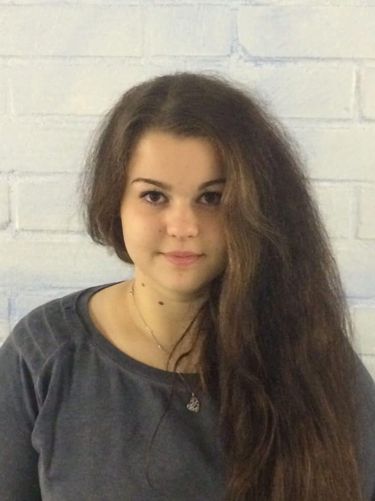 Eine junge Frau mit langen braunen Haaren und grauem Oberteil steht vor einer hellen Backsteinwand und blickt mit neutralem Gesichtsausdruck direkt in die Kamera.