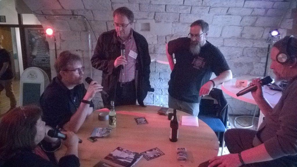 Fünf Personen sitzen und stehen um einen runden Holztisch in einem dunklen Raum mit Steinwänden und unterhalten sich über Mikrofone. Auf dem Tisch liegen Getränkeflaschen, Broschüren und Audiogeräte.