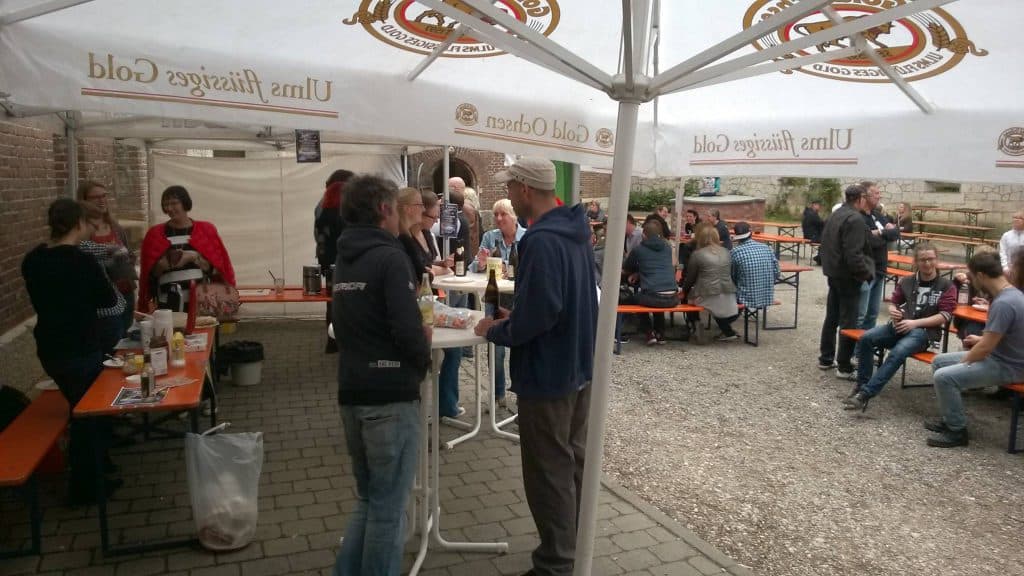 Menschen treffen sich bei einer Veranstaltung im Freien unter großen weißen Sonnenschirmen. Sie stehen und sitzen an orangefarbenen Bänken und Tischen, unterhalten sich und essen. Die Atmosphäre wirkt ungezwungen, eine Backsteinmauer und ein Kiesboden sind sichtbar.