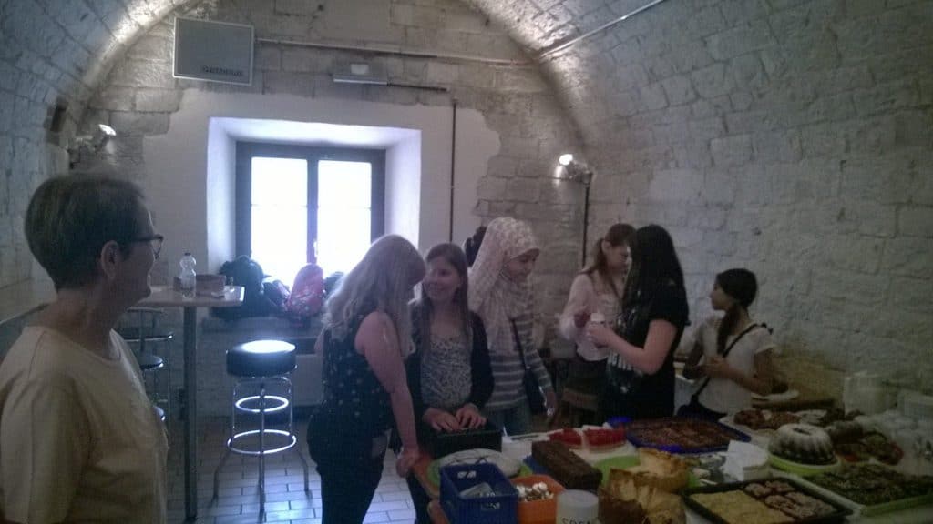 Eine Gruppe von Menschen versammelt sich um einen Tisch mit verschiedenen Backwaren in einem Raum mit Steinwänden und gewölbten Decken. Einige unterhalten sich, während andere Essen servieren. Durch ein Fenster im Hintergrund fällt Tageslicht herein.