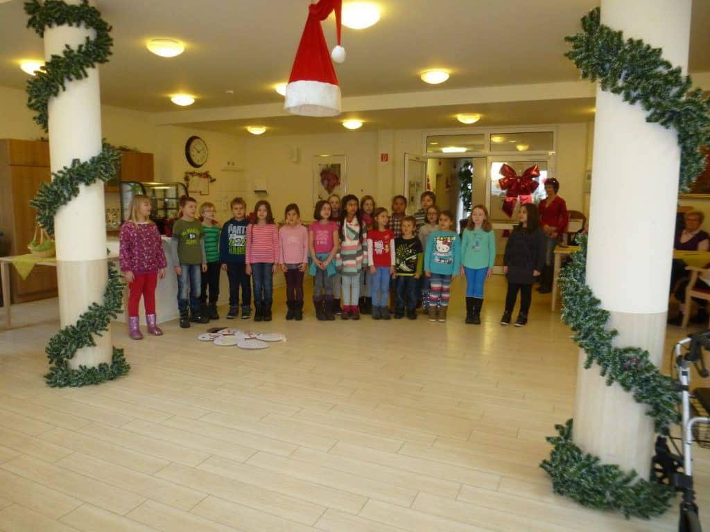 Eine Gruppe Kinder steht in einer Reihe und singt in einem dekorierten Raum mit Weihnachtsgirlanden und einer Weihnachtsmannmütze, die von der Decke hängt. Im Hintergrund sitzen Menschen, und festliche Dekorationen schmücken den Raum.