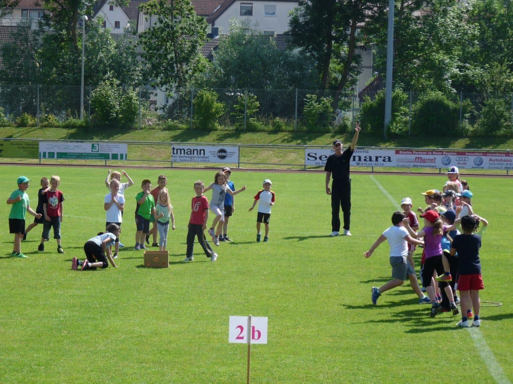 Kinder spielen und nehmen an Gruppenaktivitäten auf einem Rasensportplatz teil, wobei ein Erwachsener sie beaufsichtigt. Im Hintergrund sind Bäume und Gebäude zu sehen, im Vordergrund ein Schild mit der Aufschrift 2b.