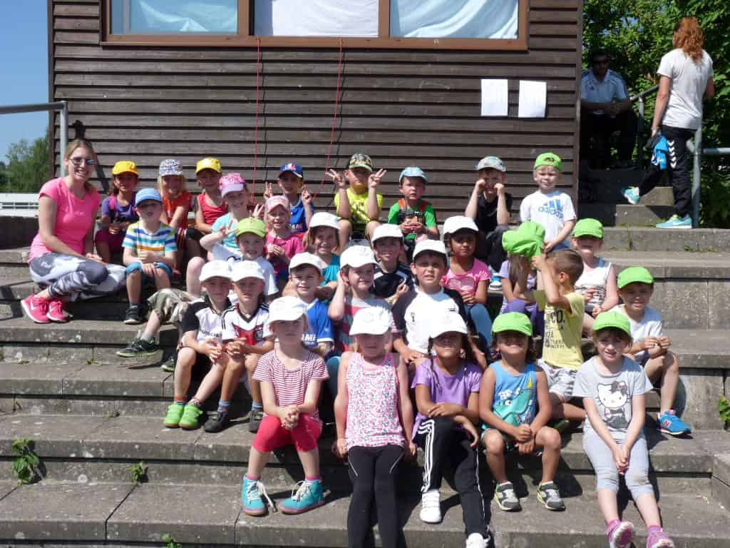 Eine Gruppe kleiner Kinder und zwei Erwachsene sitzen auf einer Außentreppe und lächeln in die Kamera. Die meisten Kinder tragen bunte Hüte und Freizeitkleidung. Hinter ihnen steht ein Holzgebäude mit Fenstern.